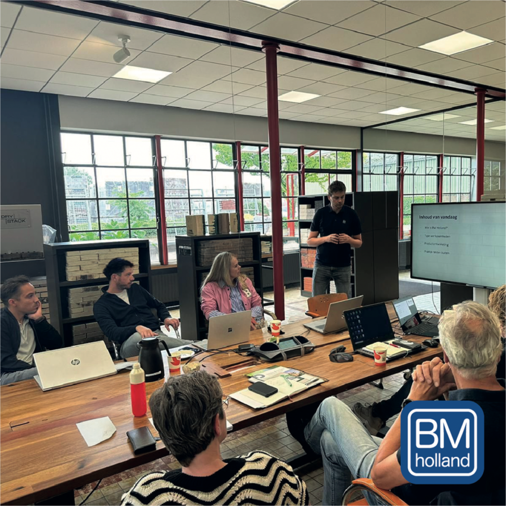 Een groep van zes mensen zit met laptops rond een tafel in een modern kantoor en kijkt naar een man die een presentatie geeft op een scherm. Door de grote ramen valt daglicht naar binnen. In de rechteronderhoek staat het logo van BM Holland.