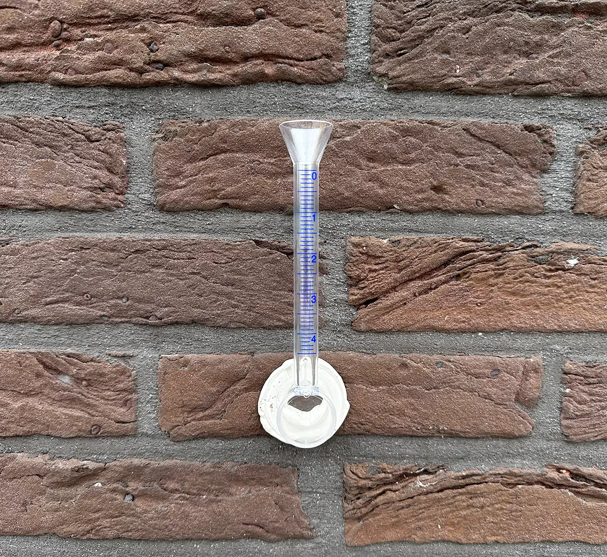 Een doorzichtige regenmeter met blauwe markeringen is met behulp van een witte zelfklevende voet aan een bakstenen muur bevestigd.