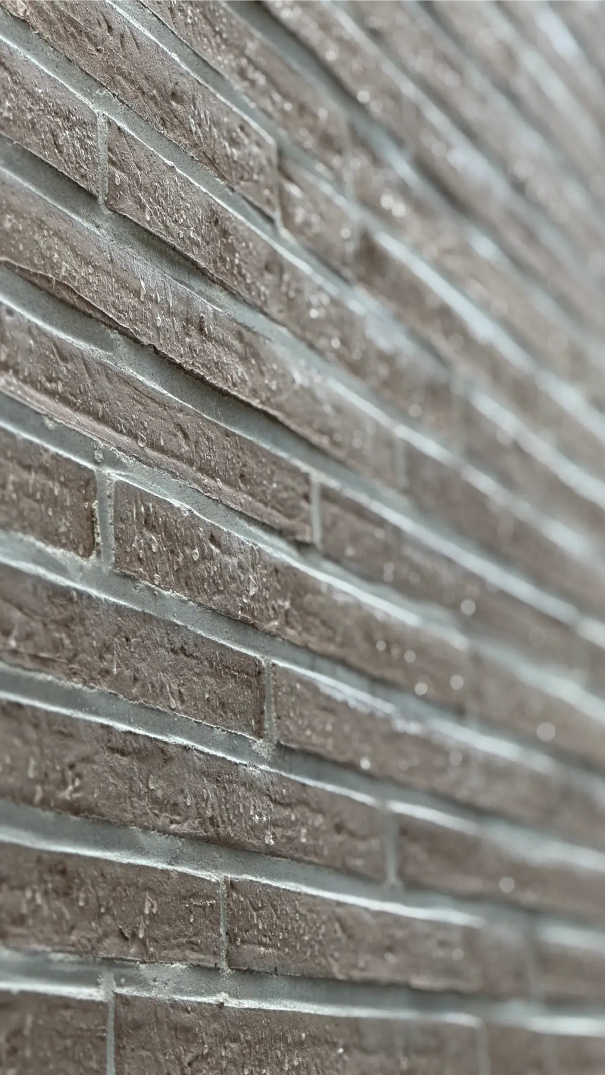 Een close-up van een bruine bakstenen muur met horizontale lijnen, waarop waterdruppels op het oppervlak te zien zijn. De foto is vanuit een schuine hoek genomen, waardoor een gevoel van diepte en perspectief ontstaat.