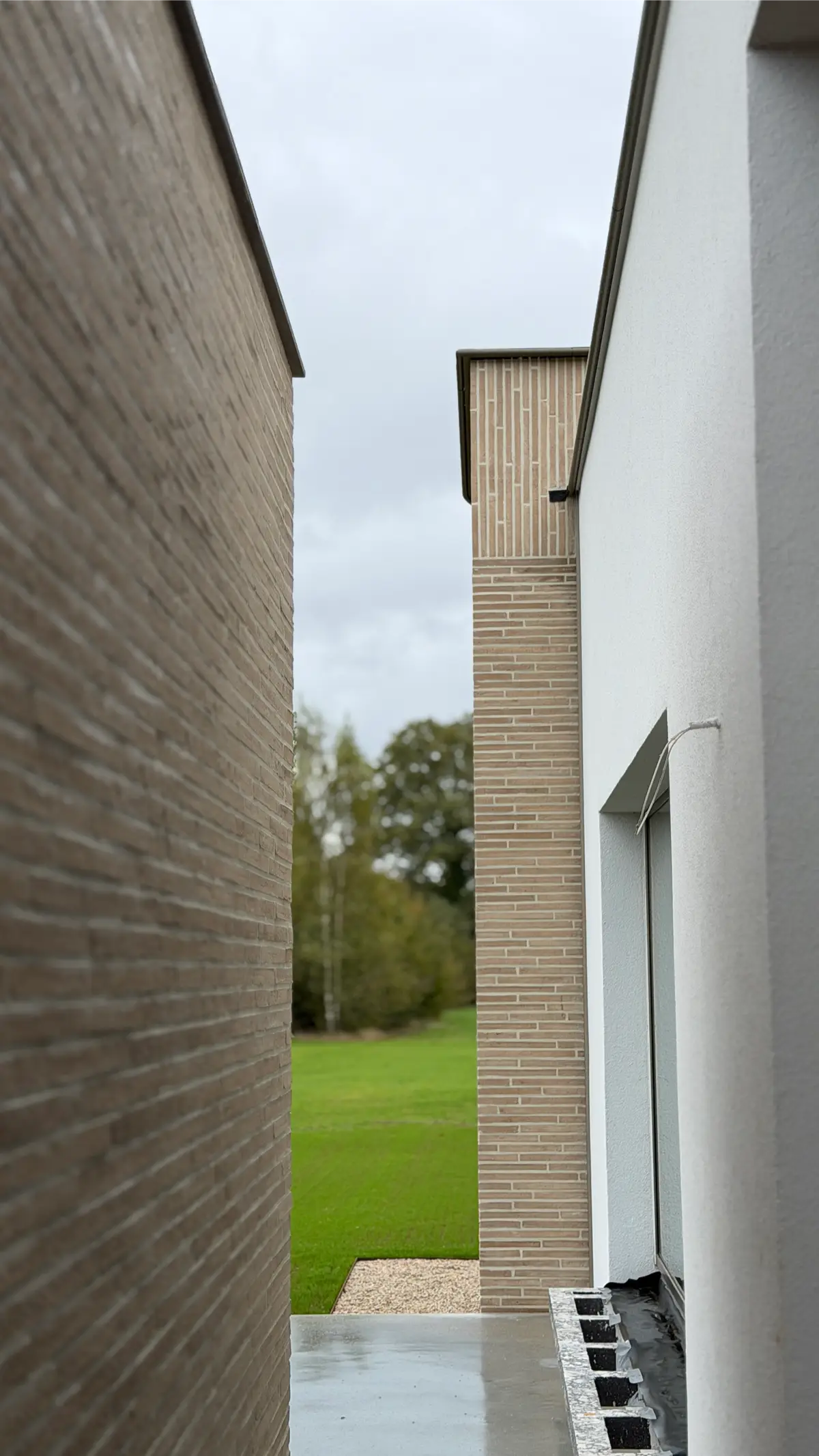 Een smal uitzicht tussen twee moderne gebouwen met beige en witte muren, met op de achtergrond groen gras, bomen en een bewolkte hemel.
