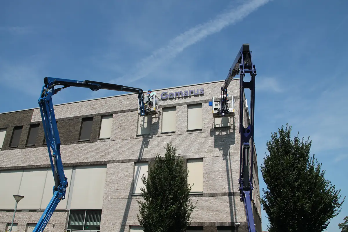 Twee werknemers op hoogwerkers zijn bezig met het plaatsen of repareren van een bord met het opschrift „Compus“ op de bovenste gevel van een modern bakstenen gebouw, onder een helderblauwe hemel. Op de voorgrond zijn bomen te zien.