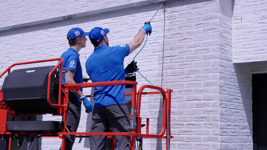 Twee werknemers in blauwe uniformen staan op een rode hoogwerker en maken met een slang de buitenmuur van een lichtgekleurd bakstenen gebouw schoon of behandelen deze.