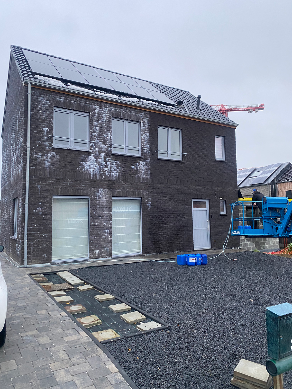Een huis van twee verdiepingen, opgetrokken uit donkere baksteen, met zonnepanelen op het dak. Op de bakstenen is een witte aanslag zichtbaar. In de tuin staan een blauwe lift, bouwmaterialen en is de tuin nog niet helemaal aangelegd.