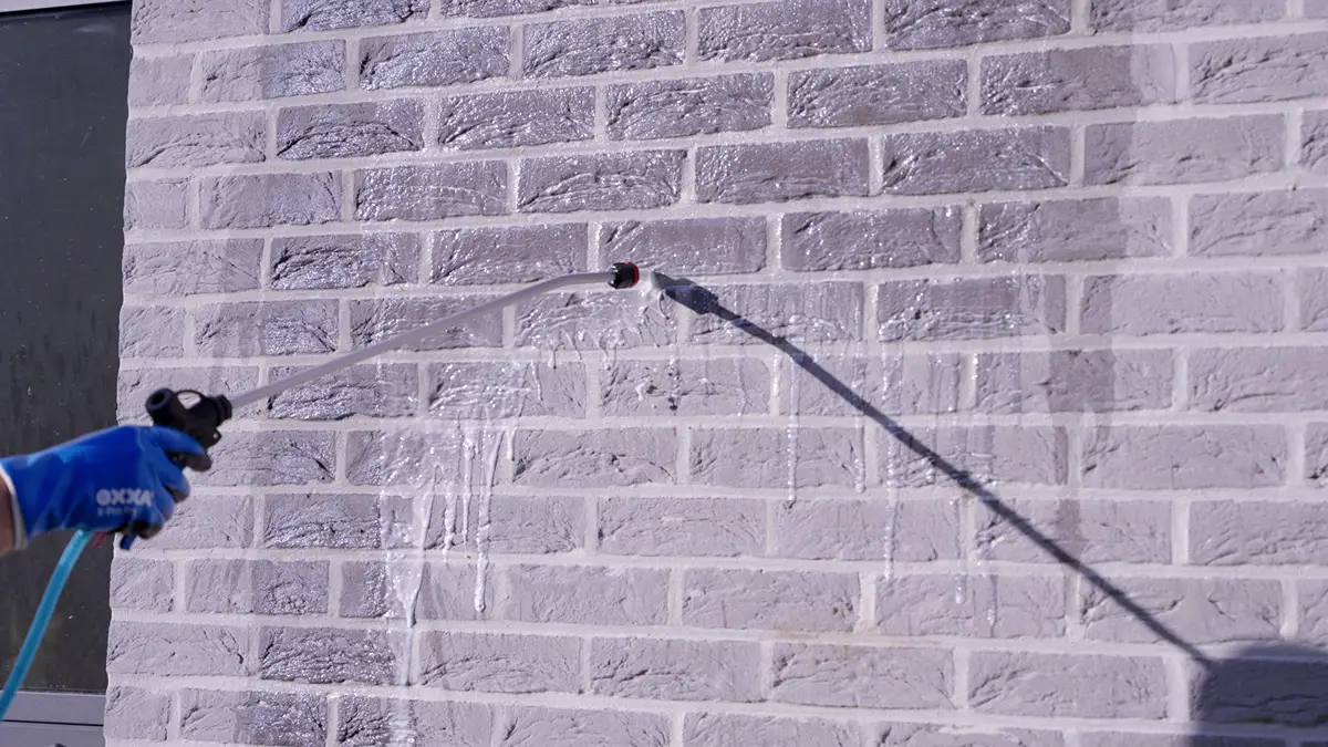 Iemand met een blauwe handschoen sproeit met een tuinslang water op een lichtgrijze bakstenen muur, waarbij het water via een gat in de muur langs de stenen naar beneden stroomt.