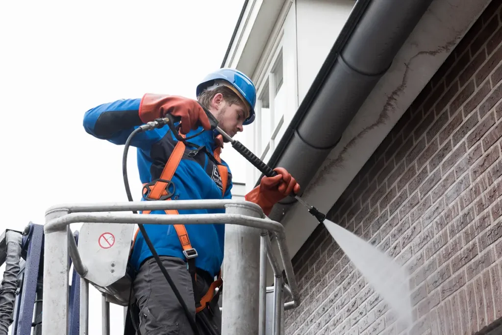 Een werknemer in veiligheidsuitrusting staat op een hoogwerker en maakt met een hogedrukreiniger de bakstenen muur en de dakgoten van een gebouw schoon.