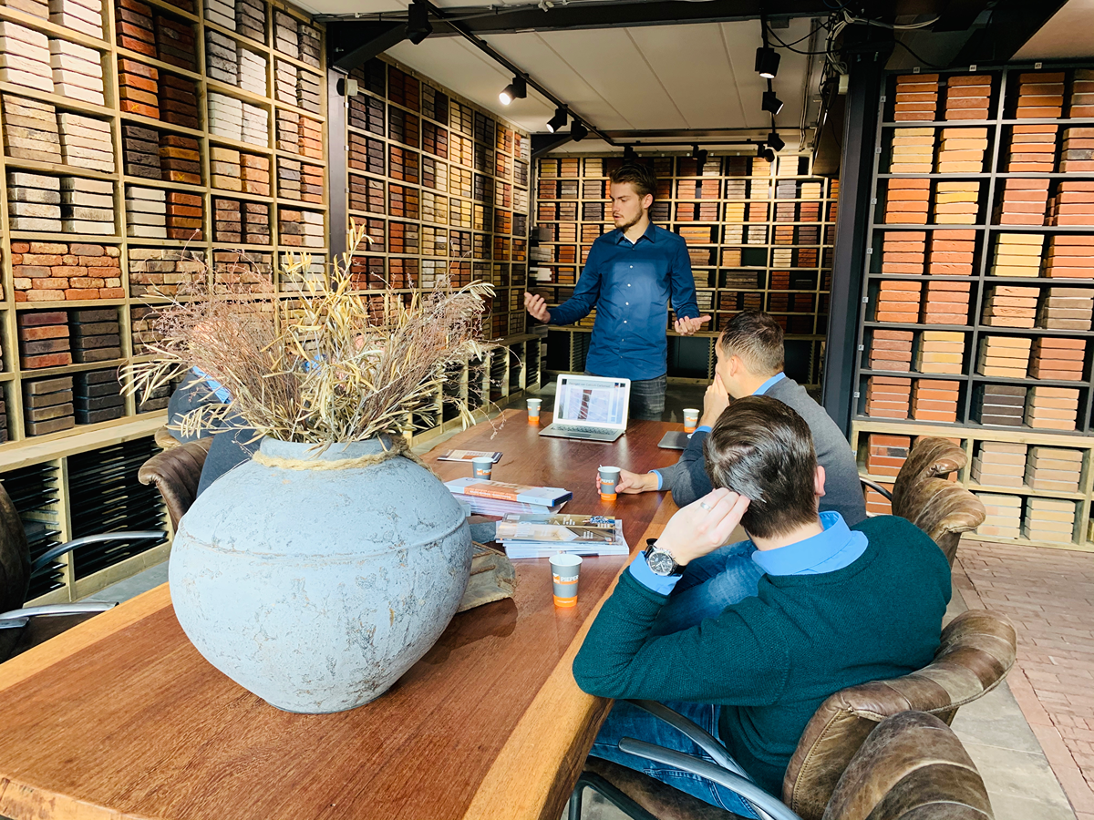 Een man staat een presentatie te geven aan drie mensen die rond een tafel zitten in een kamer waar overal voorbeelden van bakstenen staan uitgestald. Op de tafel staan een laptop, documenten en koffiekopjes, samen met een grote vaas met gedroogd gras.