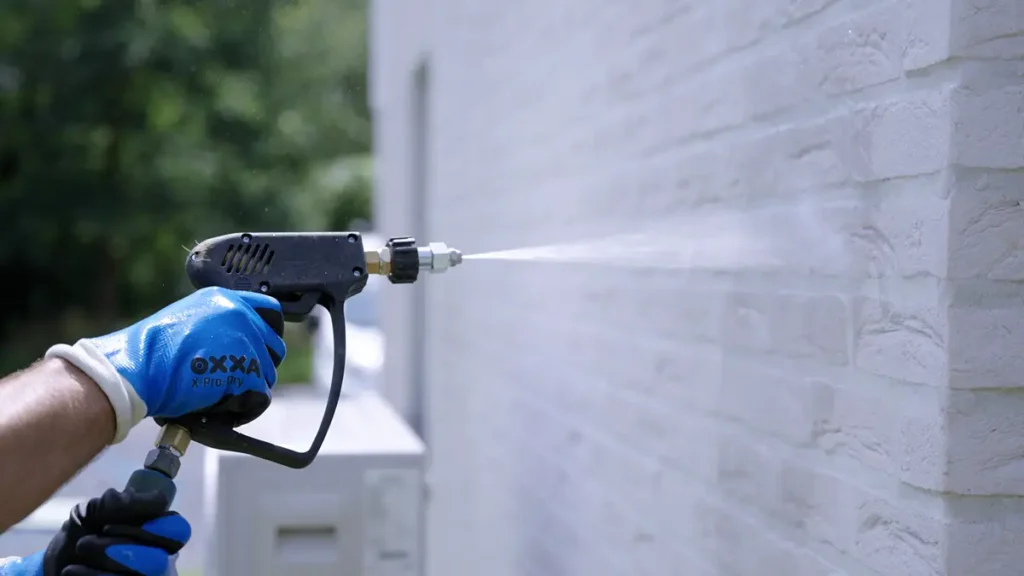 Iemand met blauwe handschoenen aan maakt met een hogedrukreiniger een witte bakstenen muur buiten schoon of spuit er water op.