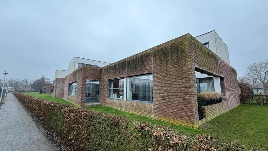 Een modern, gelijkvloers bakstenen gebouw met grote ramen en platte daken staat naast een trottoir. Het gebouw wordt omringd door een bewolkte hemel, geknipte heggen en een grasveld.