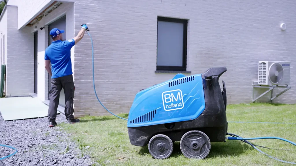 Iemand gebruikt een blauwe hogedrukreiniger met het opschrift „BM Holland“ om de buitenmuur van een lichtgekleurd gebouw schoon te maken. De hogedrukreiniger staat op het gras ernaast.