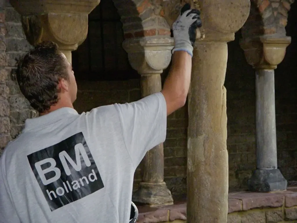 Een arbeider die een BM Holland-shirt en handschoenen draagt, is bezig met het schoonmaken of restaureren van een stenen zuil in een historisch gebouw, met op de achtergrond gewelfd metselwerk en zuilen.