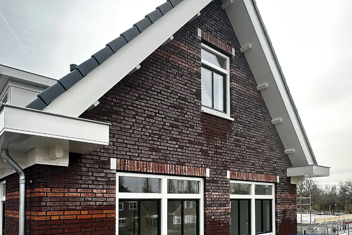 Een huis van rode baksteen met ramen met witte kozijnen, een steil grijs dak en zichtbare witte dakranden. De lucht is bewolkt en op de achtergrond is een deel van een steiger te zien.
