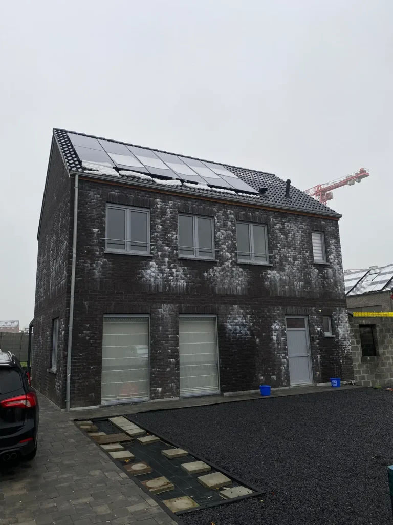 Een huis van twee verdiepingen, opgetrokken uit donkere baksteen, met witte vlekken op de muren en zonnepanelen op het dak; links staat een geparkeerde auto en op de achtergrond is op een bewolkte dag een bouwkraan te zien.