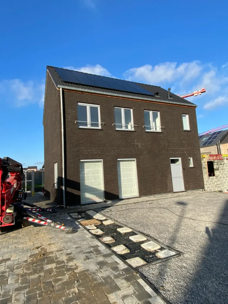 Een moderne woning met twee verdiepingen, een donkere bakstenen gevel, drie hoge ramen, zonnepanelen op het dak en een geplaveide oprit onder een strakblauwe hemel. Aan de linkerkant is bouwmaterieel te zien.