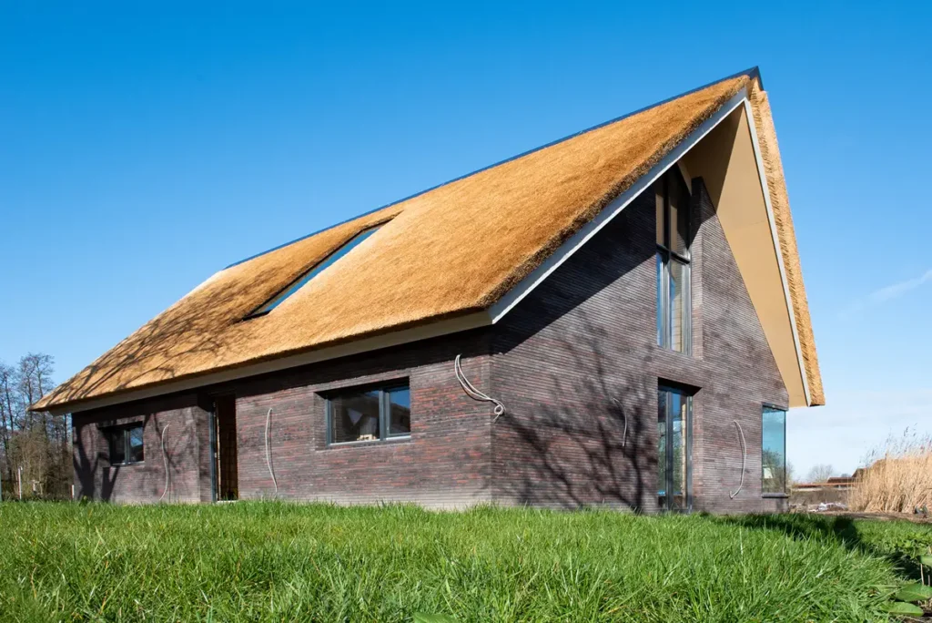 Een modern huis met donkere bakstenen muren en een steil hellend rieten dak, grote ramen, gelegen op een grasveld onder een helderblauwe hemel.