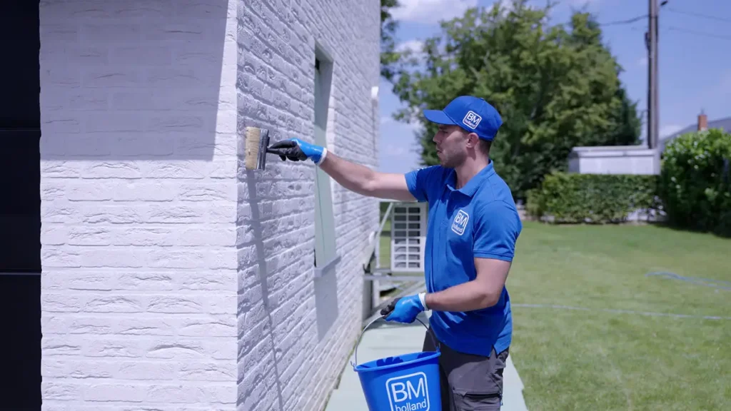 Een man in blauwe werkkleding schildert buiten een witte bakstenen muur; in de ene hand houdt hij een kwast vast en in de andere een blauwe emmer met het opschrift ‘BM Holland’. Op de achtergrond zijn een groen gazon en bomen te zien.