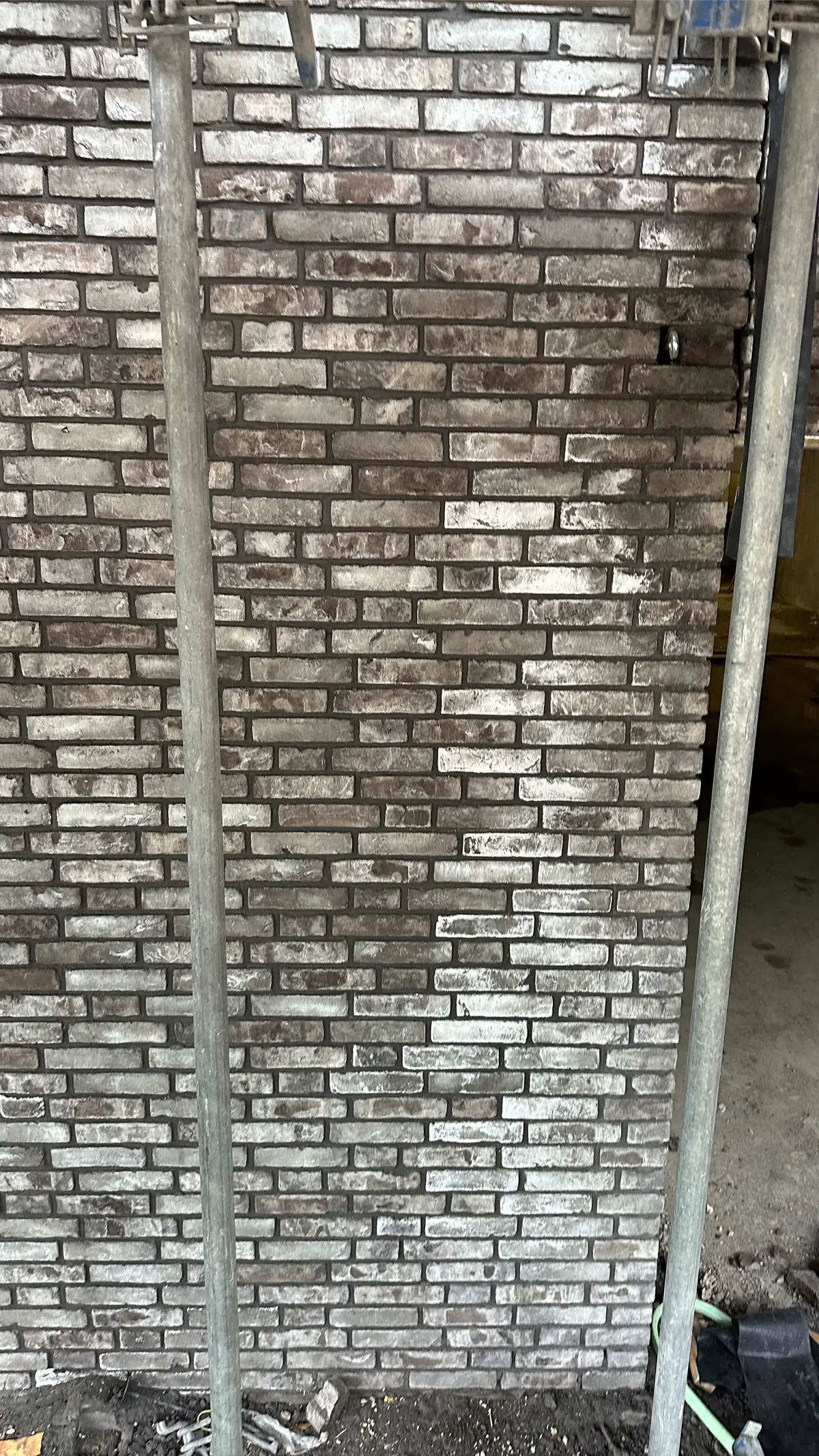 Een bakstenen muur met een verweerde uitstraling, geflankeerd door twee verticale metalen palen op de voorgrond, en hier en daar wat bouwmaterialen onderaan.