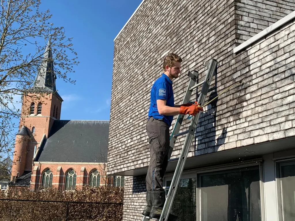 Een man die op een ladder staat, reinigt de dakspanen van een modern gebouw met een hogedrukreiniger. Op de achtergrond is onder een helderblauwe hemel een kerk met een hoge toren te zien.