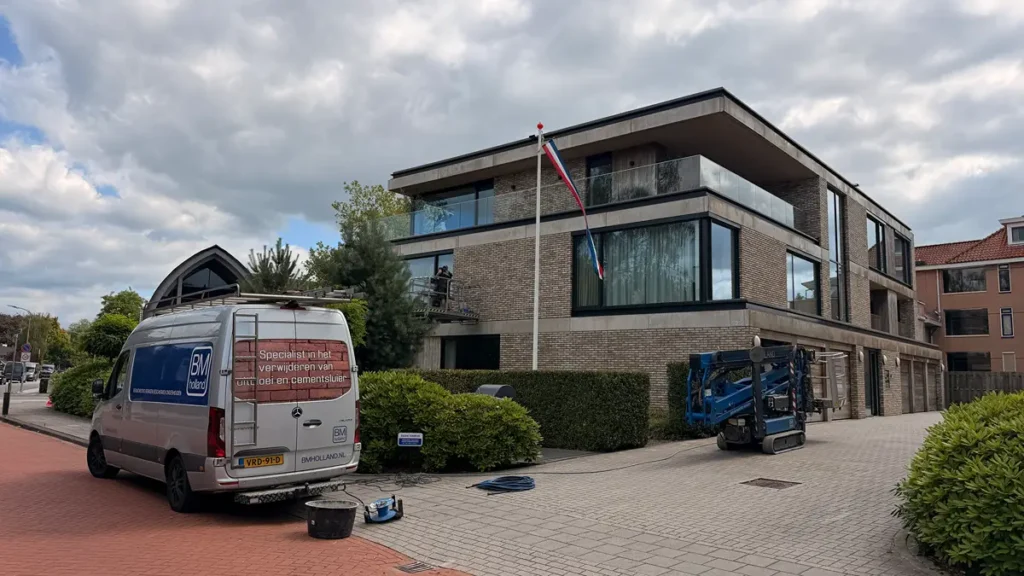 Een modern appartementencomplex met glazen balkons en een Nederlandse vlag aan een vlaggenmast buiten. Vlakbij staan een wit bestelbusje en een blauwe lift geparkeerd, en op de grond naast een heg ligt wat materieel. De lucht is gedeeltelijk bewolkt.