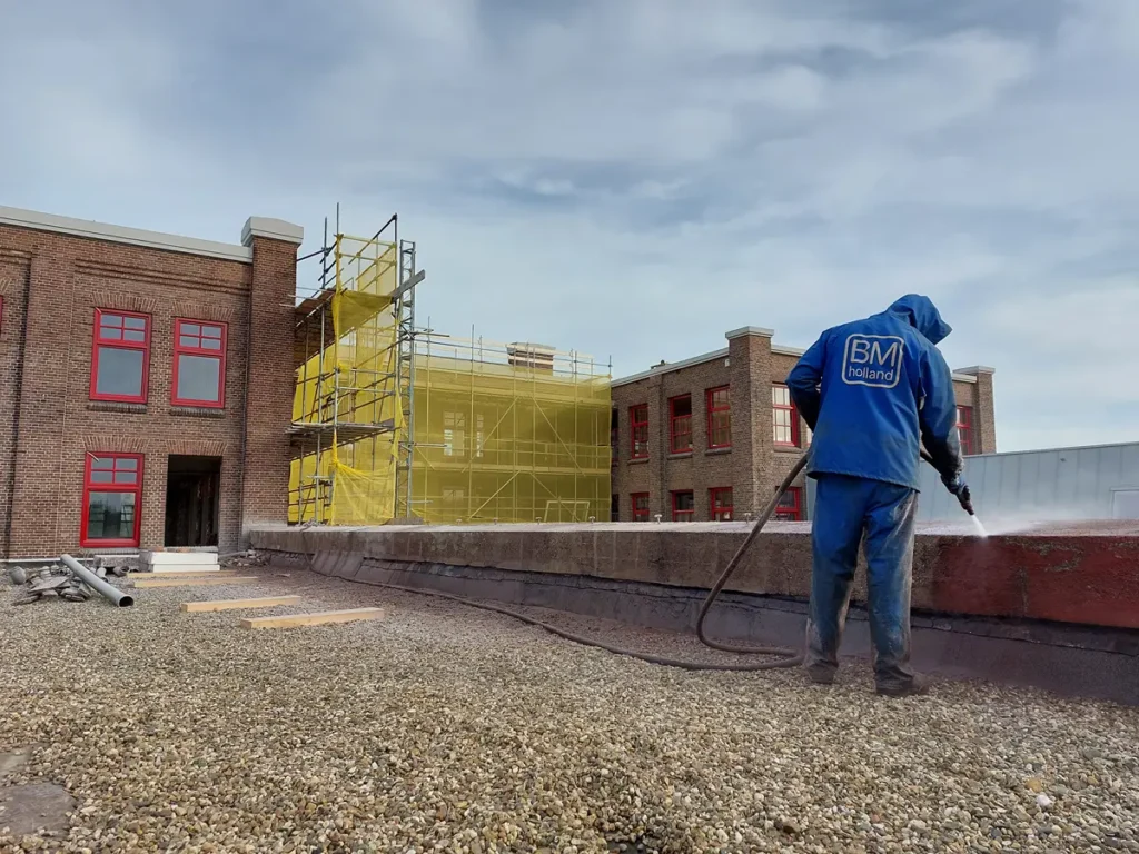 Een werknemer in blauwe beschermende kleding reinigt een dakrand met een hogedrukreiniger. Op de achtergrond staat een bakstenen gebouw met rode ramen en gele steigers, onder een gedeeltelijk bewolkte hemel.
