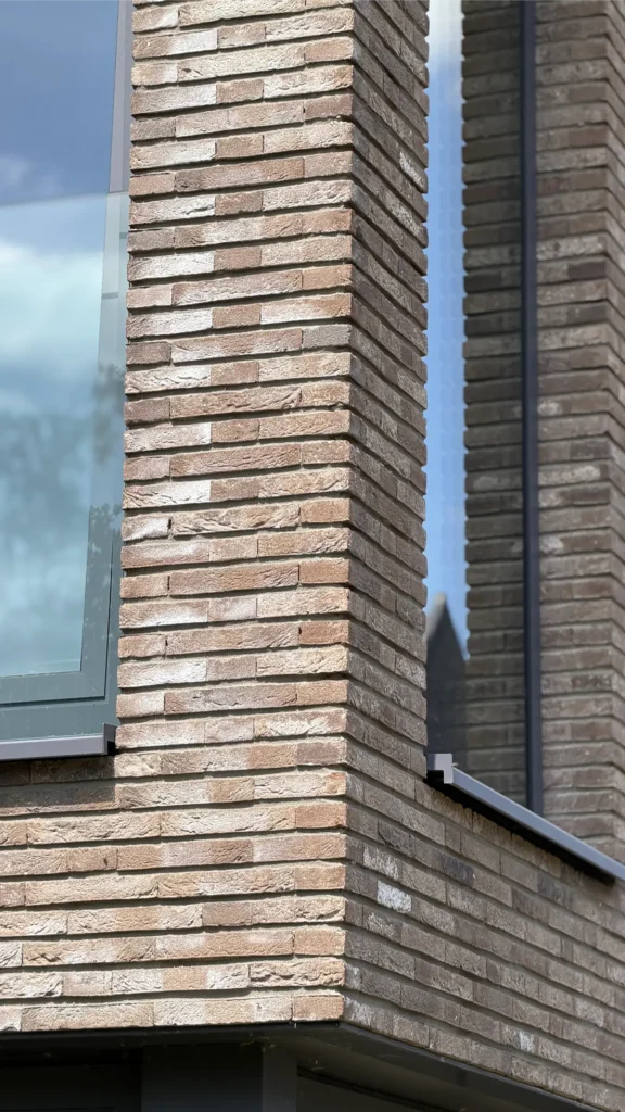 Een close-up van de hoek van een modern gebouw met lichtbruine bakstenen muren, een groot raam aan de linkerkant en weerspiegelingen van de lucht en de bomen in het glas.