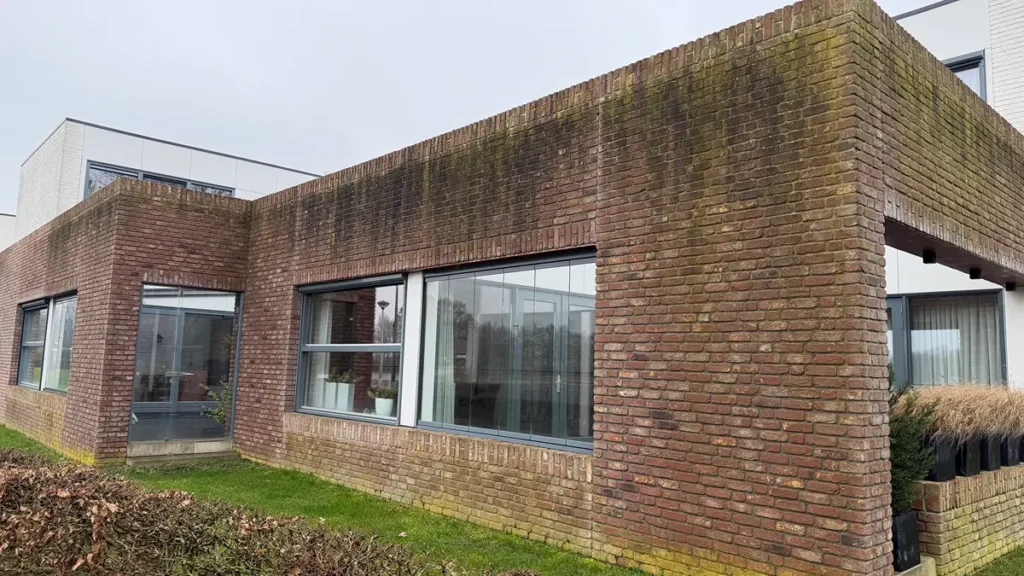 Een modern bakstenen gebouw met grote ramen, waarop aan de bovenkant van de buitenmuren groen mos of schimmel te zien is. Het gebouw wordt omzoomd door gras en gesnoeide heggen. De lucht is bewolkt.