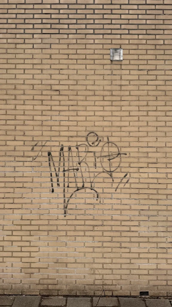 Een bruine bakstenen muur met graffiti waarop in zwarte spuitverf ‘MARIE’ lijkt te staan. Boven de graffiti bevindt zich een klein ventilatierooster en onderaan de afbeelding is de stoep te zien.