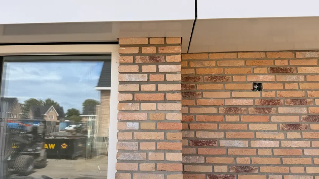 Een close-up van de buitenkant van een gebouw, waarop te zien is dat het metselwerk tussen twee delen niet op elkaar aansluit, met links een raam en rechts op de muur een klein zwart voorwerp.