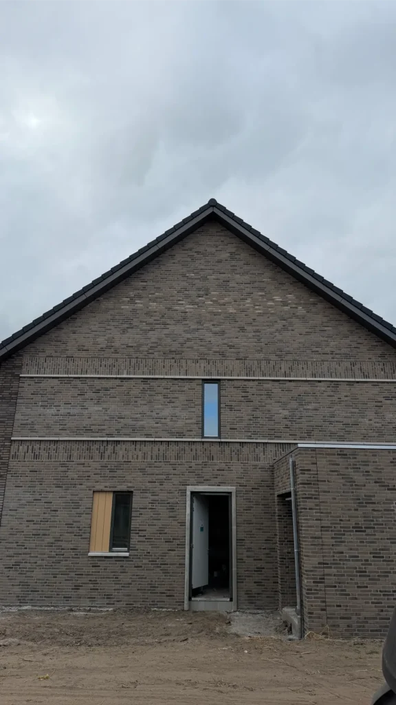 Een moderne bakstenen woning met een puntdak, een smal verticaal raam boven de deur, een overdekte ingang en een dichtgetimmerd raam aan de linkerkant. De lucht is bewolkt.