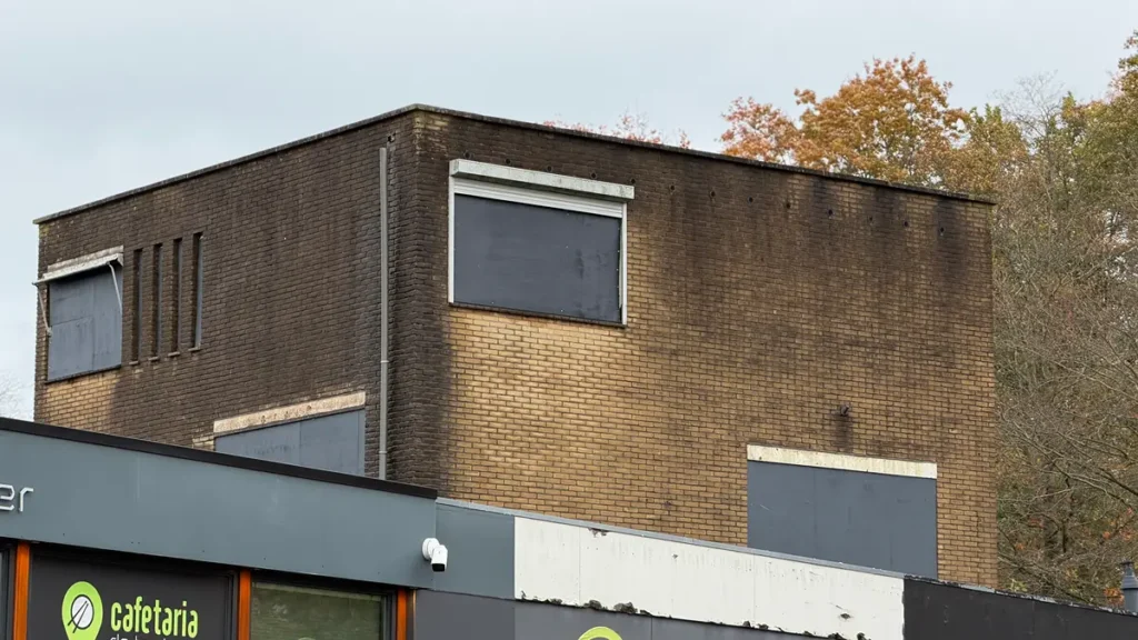 Een bakstenen gebouw met donkere, door roet bevlekte bovenste muren en dichtgetimmerde ramen torent uit boven een modern gebouw met uithangborden van een cafetaria. Op de achtergrond zijn bomen met herfstbladeren te zien.