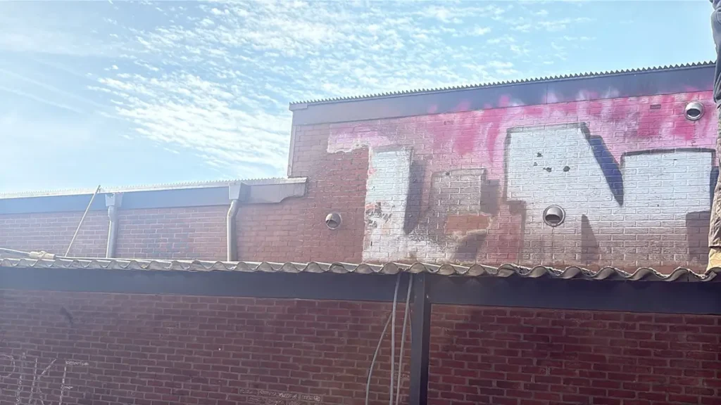 Een bakstenen gebouw met vervaagde roze en witte graffiti, gezien tegen een blauwe lucht met hier en daar wat wolken. Het dak is bedekt met golfplaten en op de voorgrond is een schaduw te zien die door een overhang wordt geworpen.