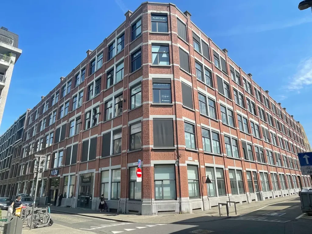 Een groot kantoorgebouw van rode baksteen met veel ramen op een zonnige dag, gelegen op een straathoek waar enkele geparkeerde auto’s en fietsen staan en een blauw eenrichtingsbord te zien is.