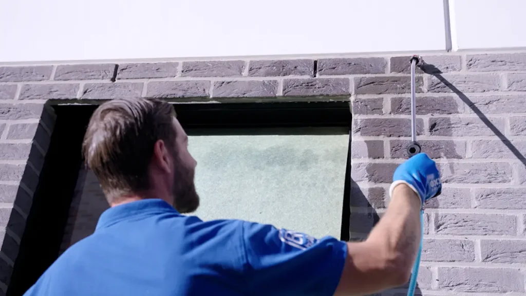 Iemand in een blauw shirt en handschoenen brengt met een spuitbus een middel aan langs de bovenrand van een bakstenen muur bij een raam.